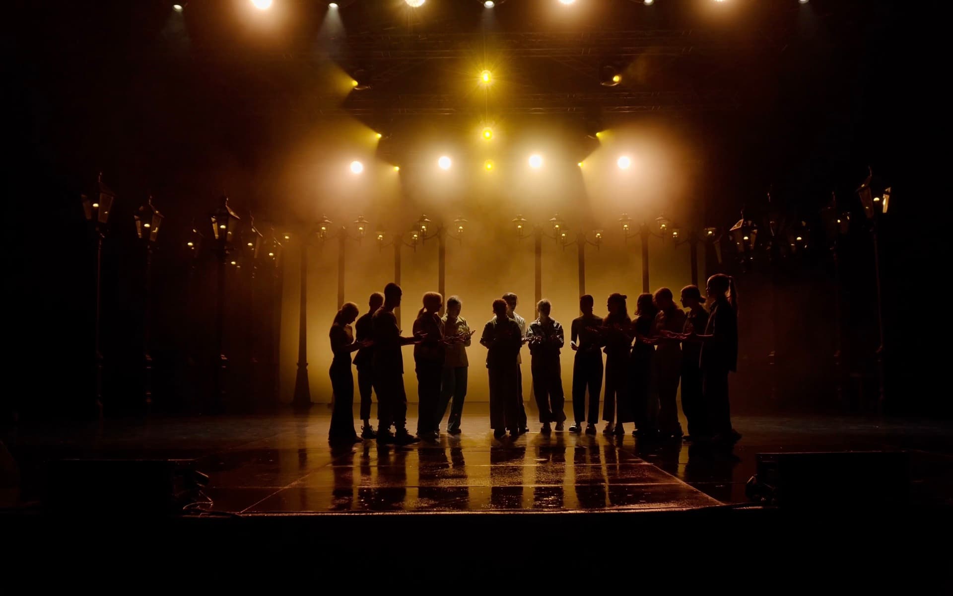 Fourteen performers standing together in silhouette beneath warm amber stage lights at Green Theatre.